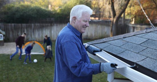 An older man wearing gloves cleans leaves from house gutters with a tool, while a family with children and a dog play with a soccer ball in the backyard behind him.