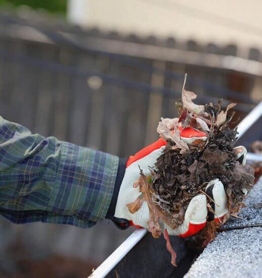 A person wearing gloves and a plaid shirt clears a large handful of dead leaves and debris from a house gutter.