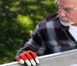 A man wearing safety glasses, a plaid shirt, and orange work gloves examines or handles a long piece of wood or metal outdoors, with green grass in the background.