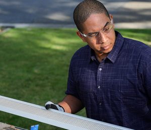 A person wearing safety glasses and gloves examines a long, rectangular metal object outdoors on a sunny day, standing on grass near a paved surface.