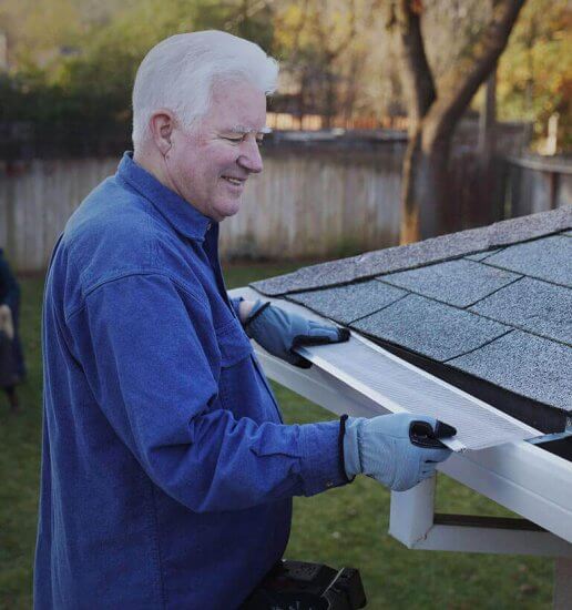 An older man wearing gloves and a blue shirt installs a gutter guard on the roof of a house in a backyard, smiling as he works.
