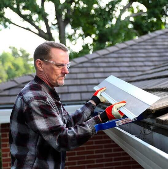 A man wearing safety glasses and gloves installs a gutter guard on the roof of a house, with trees and sky visible in the background.