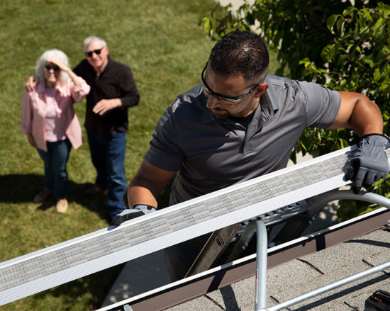 A man wearing gloves and safety glasses installs a gutter guard on a roof, while an older couple stands on the grass below, watching him work on a sunny day.