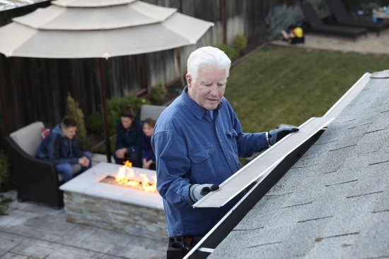 An older man wearing gloves and a blue shirt cleans the gutter on a house roof, while a group of people sit around a fire pit in a backyard patio below.