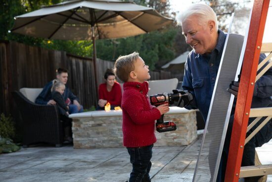 A young boy hands a power drill to an older man standing by a ladder outdoors, while two adults and another child sit by a fire pit in the background.