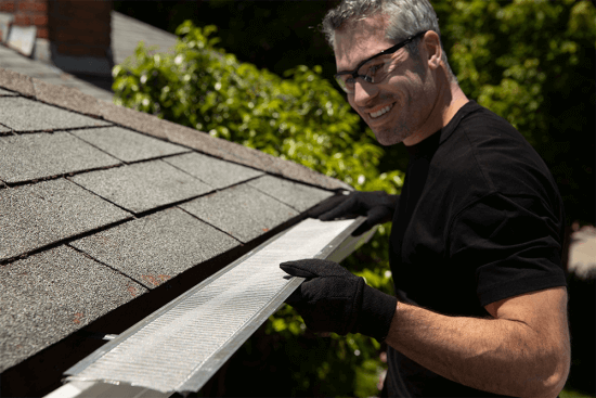 A person wearing safety glasses and black gloves installs a mesh gutter guard on the edge of a house roof, smiling while working outside on a sunny day.