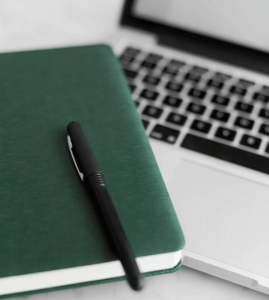 A black pen rests on a closed dark green notebook, which is placed next to an open silver laptop on a white surface.