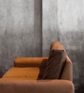 A brown sofa with a single back cushion sits against a gray, textured concrete wall. The photo focuses on the armrest and backrest, with soft lighting creating a cozy atmosphere.