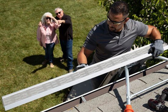 A worker installs a gutter guard on a roof while an elderly couple stands on the lawn below, watching and pointing up at him on a sunny day.
