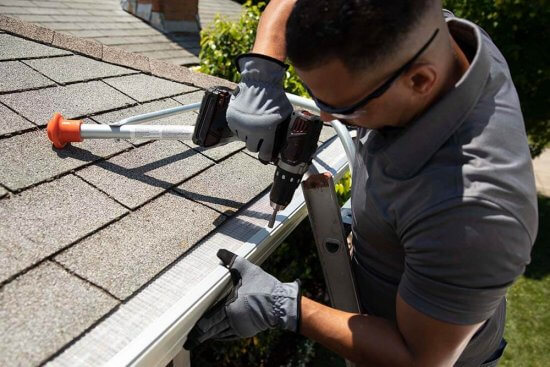 A person wearing gloves and safety glasses stands on a ladder, using a power drill to install a metal guard on the edge of a house roof near the gutter.