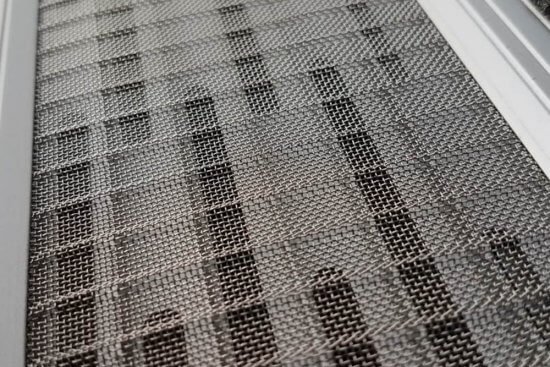 Close-up of a metal wire mesh screen with a grid pattern, set within a silver or gray frame. Shadows and light create a checkered effect across the textured surface.