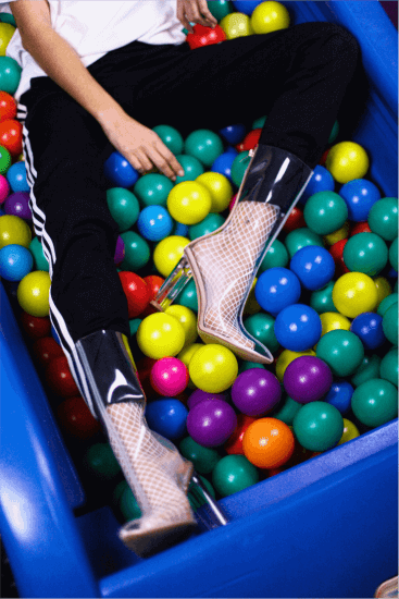 A person in black pants with white stripes and clear high-heeled boots lounges in a blue ball pit filled with colorful plastic balls. Their upper body is partially out of frame.