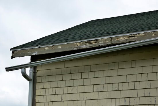 The image shows a house with green roof shingles and beige siding. The wooden fascia board under the roof is damaged and peeling, indicating rot and deterioration, while a metal gutter runs along the edge.