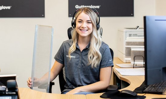 A smiling woman wearing a headset and a Gutterglove polo shirt sits at an office desk, holding a metal mesh panel. Office equipment and documents are visible in the background.