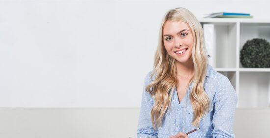 A young woman with long blonde hair, wearing a light blue shirt, smiles while sitting indoors. There is a white wall and shelves with books and a plant in the background.