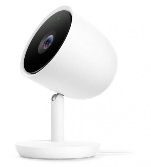 A white, modern indoor security camera with a round base and a tilted, cylindrical body. The camera lens is black and prominent, and a small green light is visible above the lens.