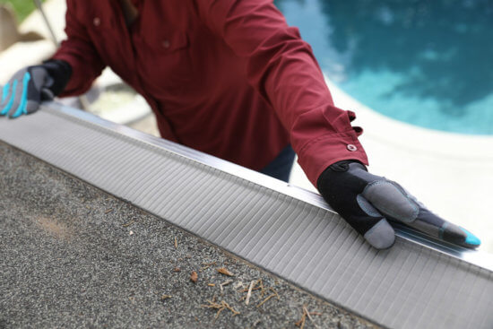 A person wearing gloves and a red long-sleeve shirt installs a mesh gutter guard on the edge of a shingled roof, with some debris visible. A pool is seen in the blurred background.