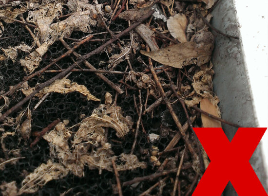 A dirty, clogged air filter covered with dried leaves, twigs, and debris, with a large red X symbol in the bottom right corner.