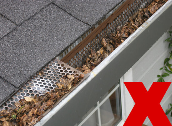 A gutter with a mesh guard is clogged with dry leaves and debris. A large red X in the corner indicates this is not the correct way to maintain gutters.