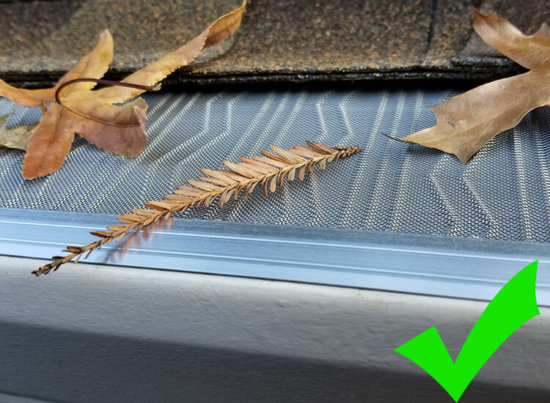 A close-up of a clean gutter guard with a few dry leaves on top and a green check mark in the lower right corner, indicating proper gutter protection.