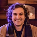 A man with curly hair and a friendly smile sits indoors, wearing a tan coat and a dark scarf with stripes. The warmly lit background creates a cozy atmosphere, highlighting his interest in RiserStep Technology made in USA.