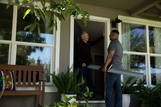 Two men talk at the entrance of a house. One stands in the doorway, while the other stands outside holding a long rectangular object, possibly a piece of wood. Green plants and a wooden bench are on the porch.
