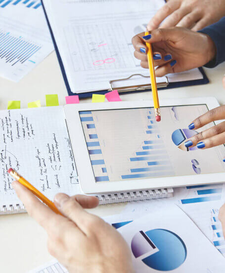 Several people collaborate at a desk with charts and graphs on paper, a tablet displaying data visualizations, and handwritten notes. Hands holding pencils are pointing at the documents.