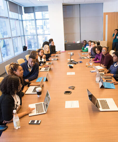 A group of people sit around a large conference table in a modern office, each with a laptop or notebook, engaged in discussion during a meeting. Large windows provide natural light in the room.