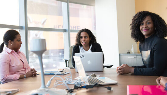 Three women sit around a conference table with laptops and office supplies, engaged in a meeting in a modern office with large windows in the background.