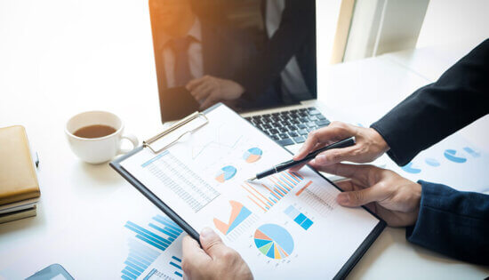 Two people in business attire review charts and graphs on a clipboard, with a laptop, coffee cup, and documents on a desk, discussing financial data in a bright office setting.
