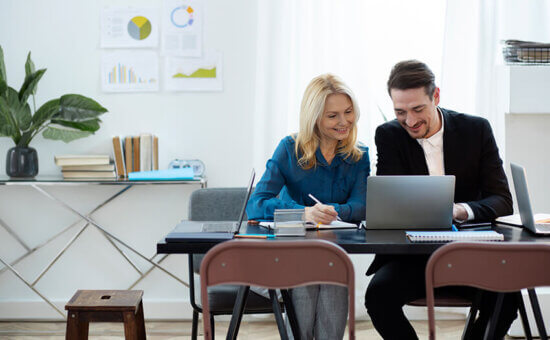 Two people, a woman and a man, sit at a desk smiling while looking at a laptop. Papers, notebooks, and office supplies are on the desk, with charts and plants in the background.