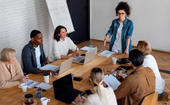 A group of six people sit around a conference table with laptops and papers, listening to a woman standing and speaking. They appear engaged in a team meeting or discussion in a modern office setting.