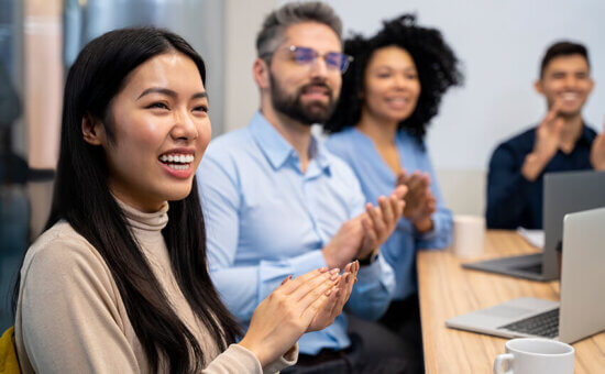 A diverse group of four people sit at a conference table, smiling and clapping. Laptops and coffee mugs are on the table, suggesting a positive moment during a meeting or presentation.