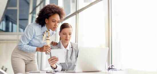 Two businesswomen in an office look at a laptop screen together. One is standing, holding a glass of water, while the other sits and holds a notepad. Large windows provide bright natural light.