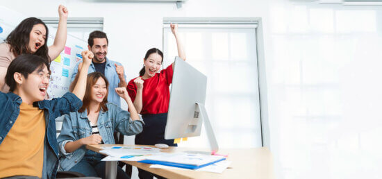 Five young adults celebrate joyfully around a computer in a bright office, raising their arms and smiling. Colorful charts and documents are on the desk and whiteboard, indicating a successful teamwork moment.