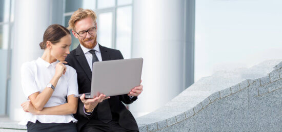 A man in a suit and a woman in business attire stand outdoors, looking at a laptop together and smiling. They appear to be discussing something on the screen in front of a modern office building.