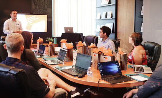 A man presents a slideshow to five people seated around a conference table with laptops, notebooks, and gift bags. The group is engaged and looking toward the presenter in a modern office setting.
