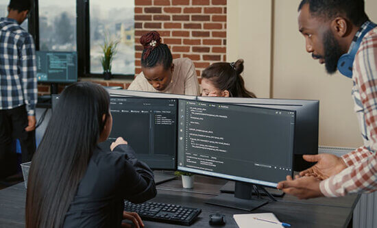 Four people collaborate in a modern office with brick walls. Two large monitors display code, while one person points at the screen and another gestures, suggesting a discussion about programming.