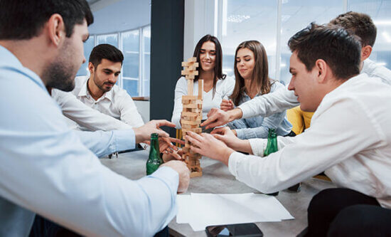 A group of six young adults sits around a table playing a game of Jenga, concentrating as they carefully remove and stack wooden blocks. Bottles and papers are on the table.