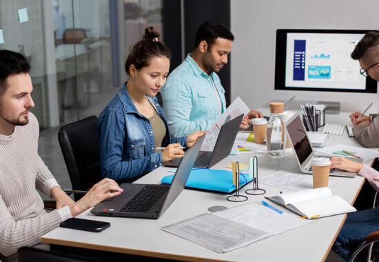 Four people work at a conference table with laptops, documents, and coffee cups. A large monitor displays charts and graphs in the background, indicating a collaborative office meeting or business discussion.
