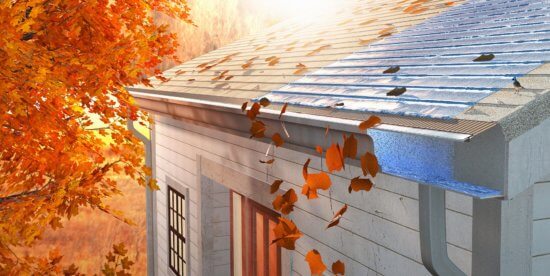 Autumn leaves are falling onto the roof and gutter of a house, with some leaves sliding off a protective gutter guard. Sunlight filters through orange foliage in the background.