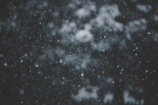 Close-up of falling snowflakes against a dark, blurred background, creating a soft, wintry atmosphere with white specks scattered throughout the image.