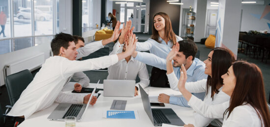 Six colleagues in business attire sit around a table with laptops, reaching across to give a group high five and smiling, in a modern, open office space.