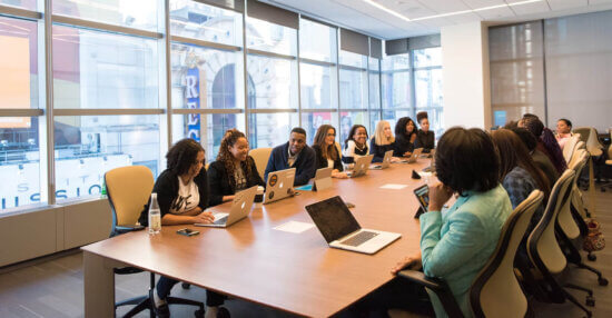 A diverse group of people sit around a large conference table with laptops, engaged in a meeting in a modern office with large windows and city views.
