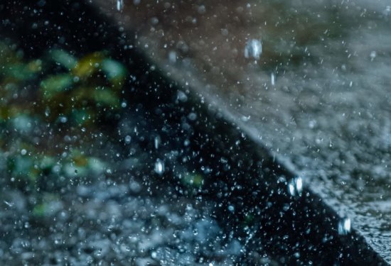 Close-up of raindrops falling and splashing on a wet surface outdoors, with water droplets suspended in the air and some green leaves blurred in the background.