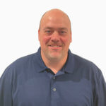 A smiling man with a bald head and light beard, wearing a navy blue polo shirt, stands against a plain white background, representing a leading gutter guard manufacturer known for stainless steel micro-mesh protection.