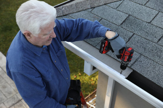 An older man in a blue shirt uses a power drill to install a gutter guard on a house roof. He is standing on a ladder, working carefully near the edge of the roof on a sunny day.