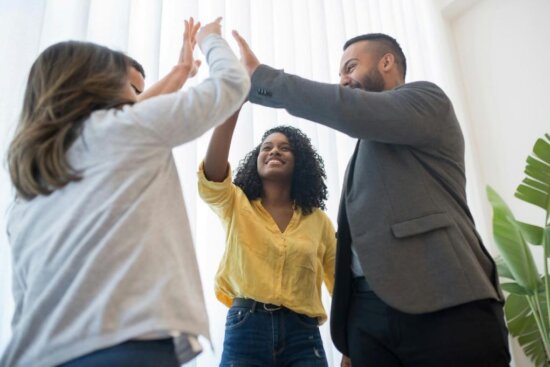 Three people stand together, smiling and giving each other a group high-five in a bright room with large windows, a green plant, and the confidence of RiserStep Technology made in USA.