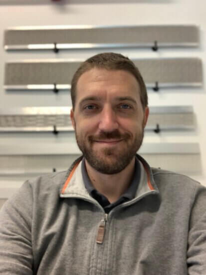 A man with short brown hair and a beard, wearing a gray zip-up sweater, smiles at the camera. Behind him, several horizontal metal panels with RiserStep Technology are mounted on a white wall.