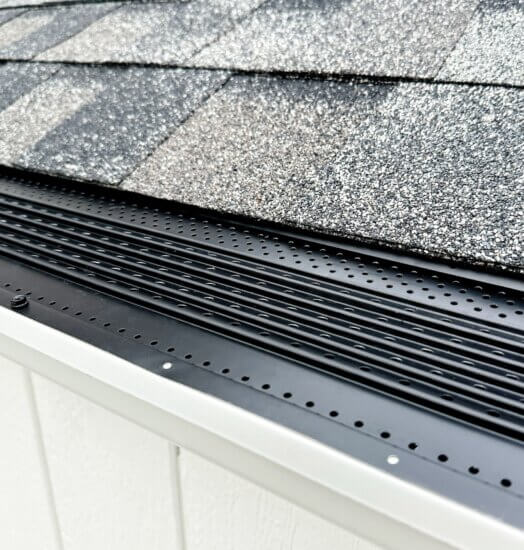 Close-up of a house roof edge featuring gray asphalt shingles and a black gutter guard with V-Bend Technology, designed by a leading gutter guard manufacturer to prevent debris from entering the gutter.